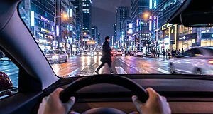 First-person view from a car at night. Hands grip the steering wheel as a pedestrian crosses a bustling, neon-lit city street with wet asphalt and traffic.