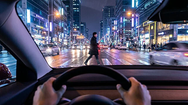 Dynamic City Nightscape from the Driver's Seat First-person view from a car at night. Hands grip the steering wheel as a pedestrian crosses a bustling, neon-lit city street with wet asphalt and traffic.