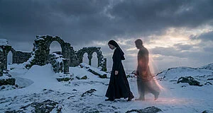 A nun walks contemplatively through a desolate, snowy landscape with ancient ruins. An ethereal, glowing figure accompanies her under a dramatic sky.