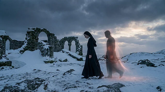 A nun walks contemplatively through a desolate, snowy landscape with ancient ruins. An ethereal, glowing figure accompanies her under a dramatic sky.