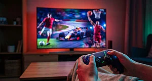 Hands hold a sleek Xbox controller with glowing accents. Blurred TV shows dynamic sports: soccer, racing, basketball. Energetic screen light.
