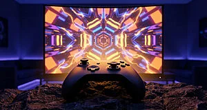 Sleek game controller on a dark rock, glowing from a large screen showing abstract, colorful, geometric retro game patterns.