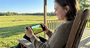 Content young adult smiles, playing a vibrant farming sim on a portable device in a wooden chair on a sunny rural porch.