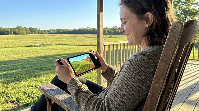 Rural Gaming Bliss Content young adult smiles, playing a vibrant farming sim on a portable device in a wooden chair on a sunny rural porch.