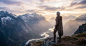 Ciri, de dos, sur une crête rocheuse, contemplant une vaste chaîne de montagnes enneigées et une rivière à l'aube.