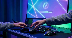 Anonymous hands on gaming controller, illuminated keyboard, and mouse. Screen shows 'V' icon, progress bar, abstract neon glow.
