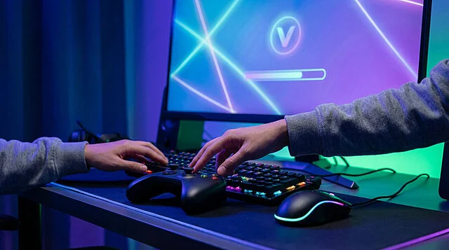Dynamic Gaming Setup: Hands, Controller, RGB Glow Anonymous hands on gaming controller, illuminated keyboard, and mouse. Screen shows 'V' icon, progress bar, abstract neon glow.