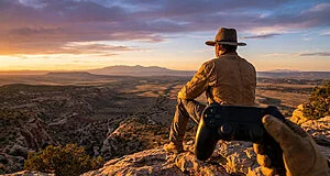Person in hat and jacket views vast western sunset landscape from rocky peak, holding a game controller in foreground.