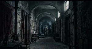 Image sombre d'un couloir ancien et délabré. Un personnage en manteau se tient dos à nous, face à un portail sombre et mystérieux.
