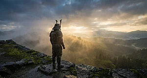A lone armored warrior stands on a mossy stone castle rampart, gazing at a misty valley illuminated by golden sun rays at dawn.