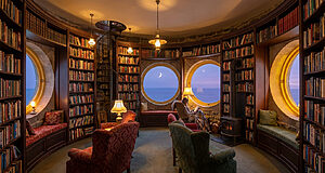 Bibliothèque circulaire cosy avec étagères de livres, fenêtres rondes sur l'océan, lune. Une personne lit près d'une cheminée.