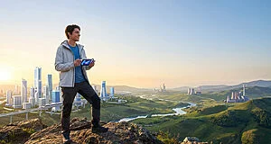 Jeune homme joue sur console au sommet d'une colline, regardant une région d'Unys mêlant ville futuriste, nature et industries.