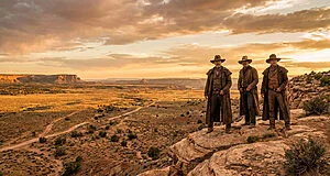 Trois hommes habillés en cow-boys, avec chapeaux et longs manteaux, se tiennent sur un rocher, dominant un vaste paysage désertique doré au coucher du soleil avec des mesas au loin.