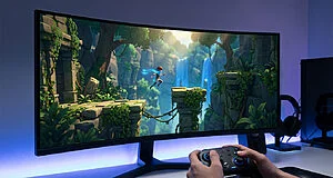 Mains tiennent une manette jouant à un jeu vidéo d'aventure sur un grand écran PC incurvé, avec un personnage sautant dans une jungle verdoyante.