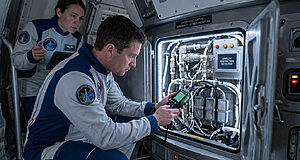 Deux astronautes examinent un panneau de système ouvert dans un vaisseau spatial. Un écran indique "AIRFLOW RESTRICTION DETECTED". L'un utilise un outil.