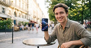 Un jeune homme souriant assis à la terrasse d'un café, tenant un smartphone affichant un fond d'écran abstrait.