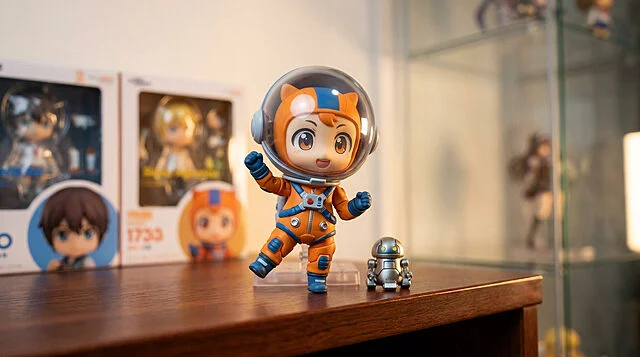 Nendoroid Esquie : La mascotte en mode astronaute Figurine Nendoroid Esquie portant une combinaison d'astronaute orange, accompagnée d'un petit robot sur une table en bois.