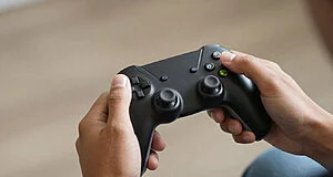 Deux mains d'une personne tenant fermement une manette de jeu vidéo noire avec des joysticks et boutons colorés sur un fond flou.