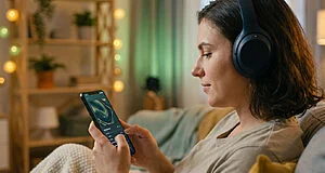 Une jeune femme avec un casque audio consulte son smartphone dans un salon chaleureux et tamisé.