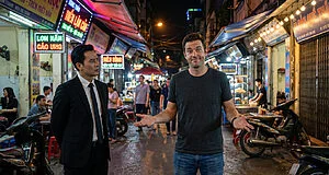 Un homme en t-shirt gris imitant le geste célèbre de Khaby Lame face à un homme en costume dans une rue vietnamienne.