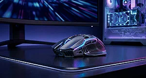 Souris gaming ergonomique grise avec éclairage LED bleu sur un tapis de bureau noir devant un PC illuminé.