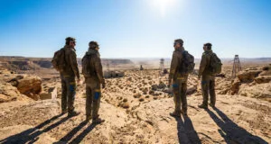 Quatre personnages en tenues de survie regardant un vaste paysage désertique et rocailleux sous un soleil éclatant.