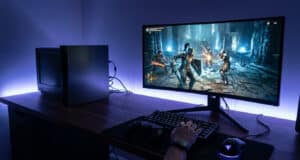 Une personne joue à un jeu vidéo sur PC avec un clavier, une souris et une console PS3 posée sur le bureau éclairé.