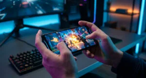 Gros plan sur des mains tenant un smartphone affichant un jeu d'action intense dans une chambre gaming éclairée en bleu.