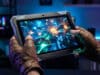Meilleures tablettes gaming 2026 : quel modèle pour jouer ? Une personne porte des gants en cuir et joue à un jeu vidéo futuriste sur une tablette robuste dans un décor gaming.