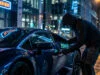 Un individu encapuchonné utilise un appareil électronique pour pirater la porte d'une voiture de sport sous la pluie.