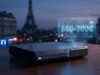 Prix de la PS6 : Sony prépare-t-il une hausse record en France ? Console de jeu futuriste projetant un hologramme avec le prix 600-700€ devant la Tour Eiffel de nuit.