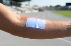 Capteur papier transpiration : l’énergie du futur pour le suivi Bras d'un athlète portant un patch en papier bleu sur la peau, sur une piste d'athlétisme en arrière-plan.