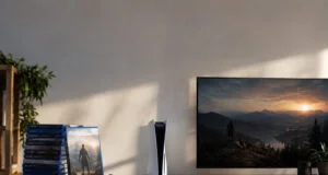 Une console PS5 et une pile de jeux physiques posées sur un meuble TV avec un écran affichant un paysage.