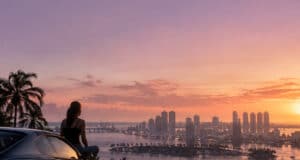 Une personne assise sur une voiture de sport, observant le coucher de soleil sur une ville côtière inspirée de Miami.