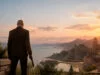 L'Agent 47 de dos, en costume, contemplant un coucher de soleil sur une ville côtière italienne avec une arme à la main.