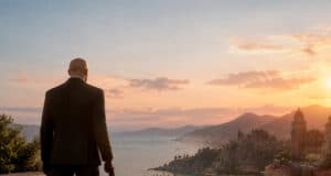 L'Agent 47 de dos, en costume, contemplant un coucher de soleil sur une ville côtière italienne avec une arme à la main.
