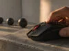 Une main utilise une souris gaming noire avec un éclairage rouge sur un socle en béton sous une lumière naturelle.
