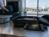 Transporter une console de jeux en avion sans la casser, mode d’emploi Une console de jeux avec un écran brisé posée sur une table en métal à côté d'une valise au contrôle de sécurité.