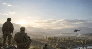 Trois soldats en tenue tactique observent une vallée montagneuse au coucher du soleil avec un hélicoptère au loin.