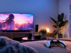 Ecchi Memories gratuit sur PS4 et PS5 : un Platine rapide Une manette PS5 posée sur un lit face à une télévision diffusant un paysage animé au coucher du soleil dans un salon.