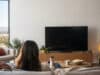 Abonnements Netflix : quel prix pour les offres en 2026 ? Une femme assise sur un canapé regarde une télévision éteinte dans un salon moderne.