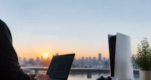 Un développeur travaille sur son ordinateur portable avec une console PS5 et une manette à ses côtés face à un coucher de soleil.