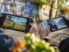 Deux mains tenant des consoles portables affichant des paysages de jeux vidéo différents sur un fond urbain.