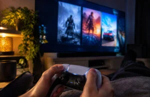 Une personne tenant une manette PlayStation 5 face à un grand écran affichant des images de jeux vidéo.