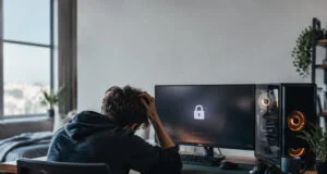 Un étudiant assis à son bureau, la tête dans les mains, face à un écran de PC gamer affichant une icône de cadenas.